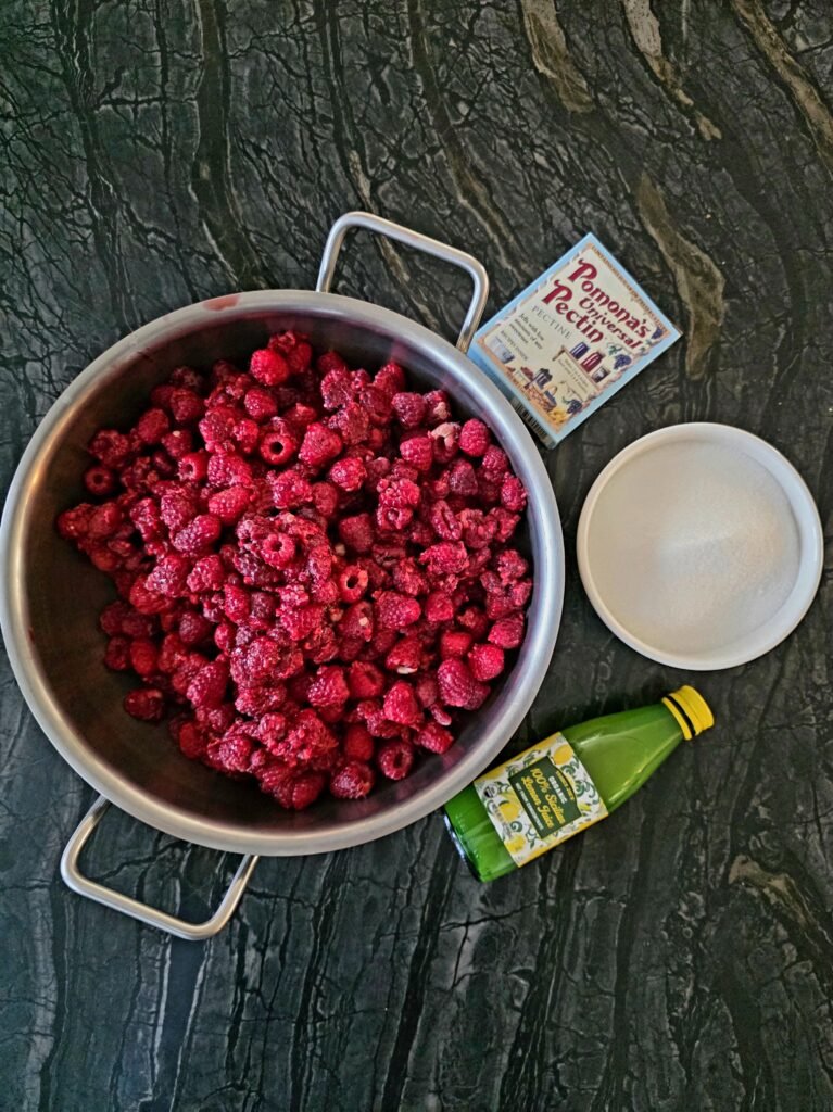 raspberries in a pot, Pomona pectin, sugar in a bowl and bottled lemon juice - ingredients used to make this jam set on a table