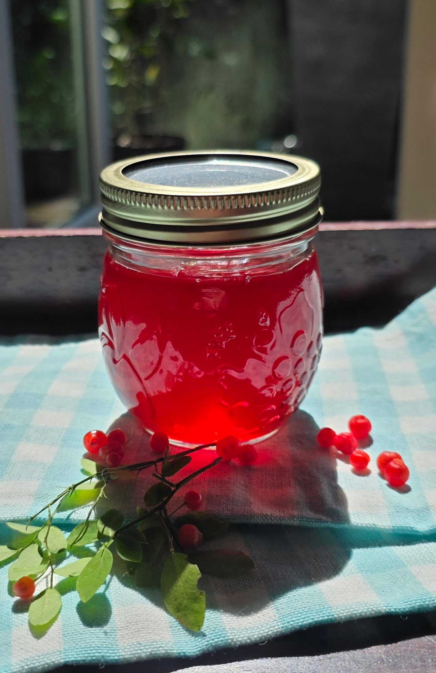 Mason jar of bright red huckleberry jelly on gingham cloth, surrounded by fresh red berries"
