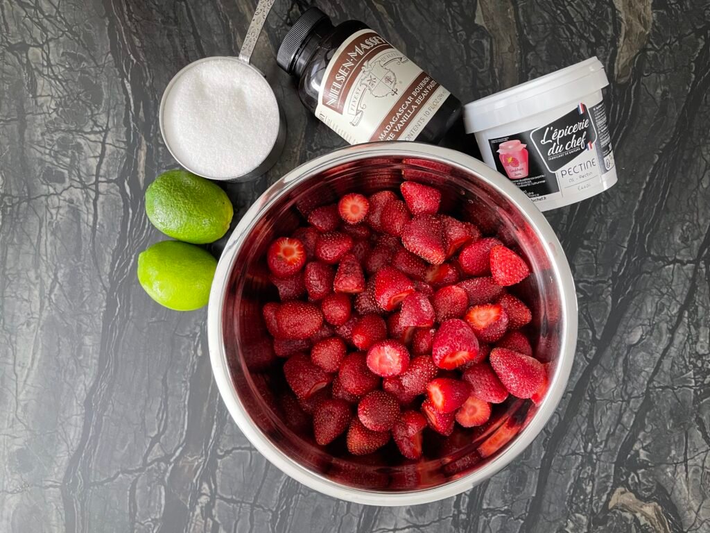 Overhead view of hulled strawberries in a stockpot surrounded by vanilla extract, vanilla paste, sugar, and a fresh lime
