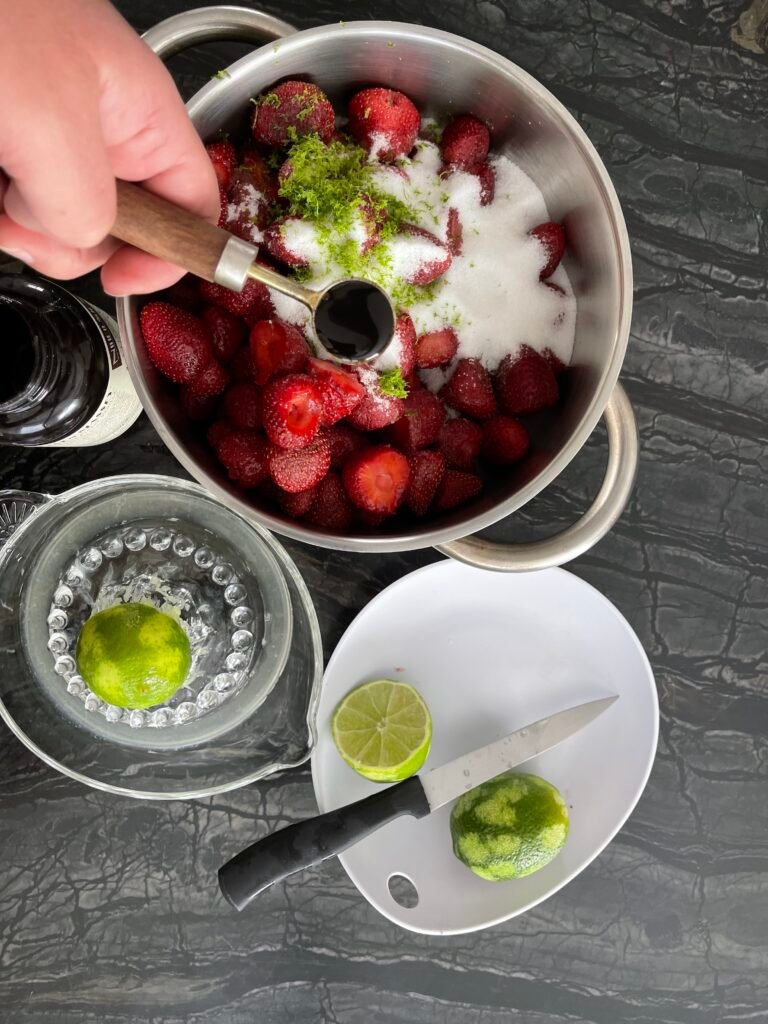 Gloved hand adding vanilla bean paste to a pot of strawberries mixed with sugar and lime zest with a plate of halved limes and a knife alongside