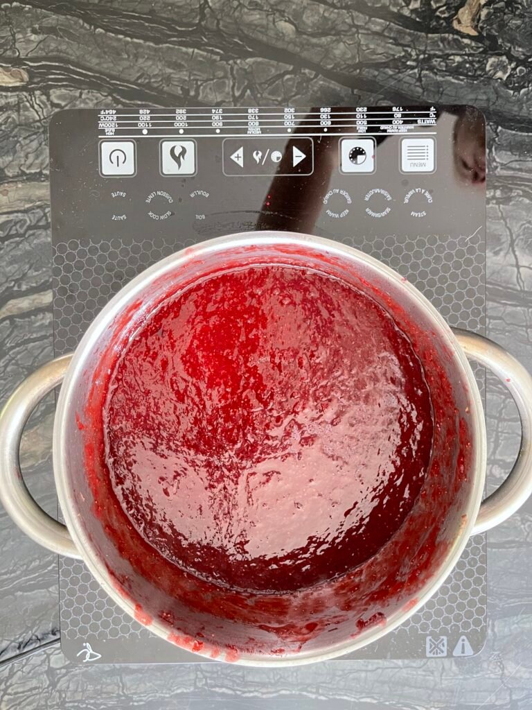Overhead view of blended strawberry lime jam cooking in a pot on the stove, glossy and deep red with a smooth, thickened surface