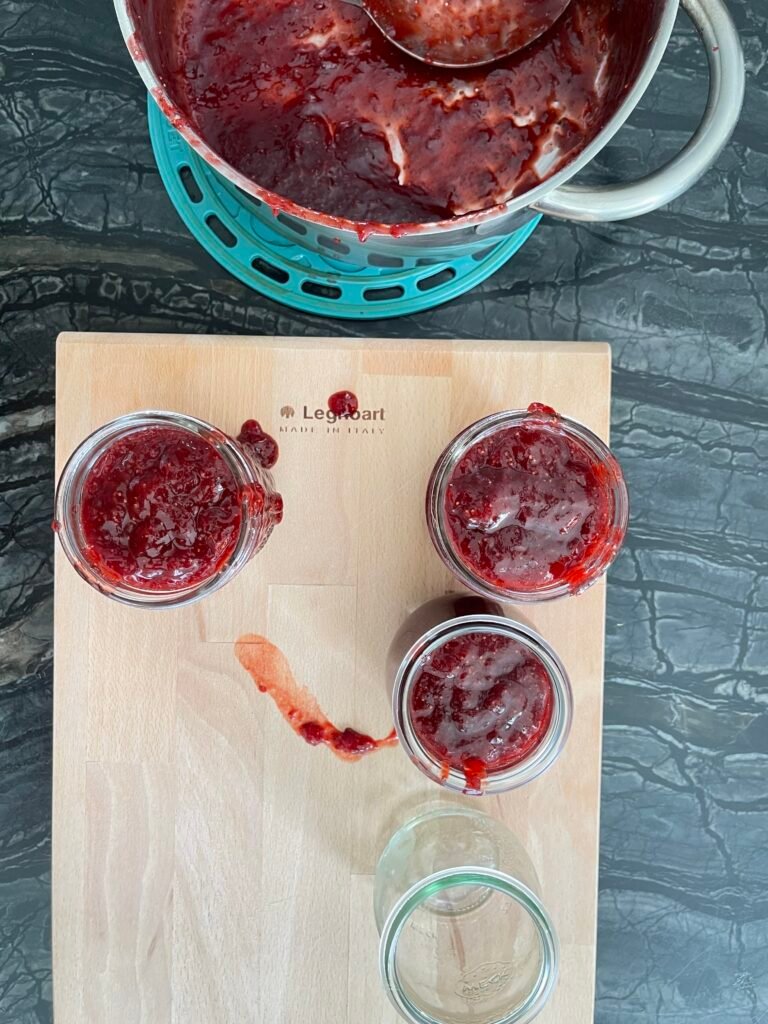 Three small canning jars partially filled with strawberry lime jam on a wooden board, with the pot in the background