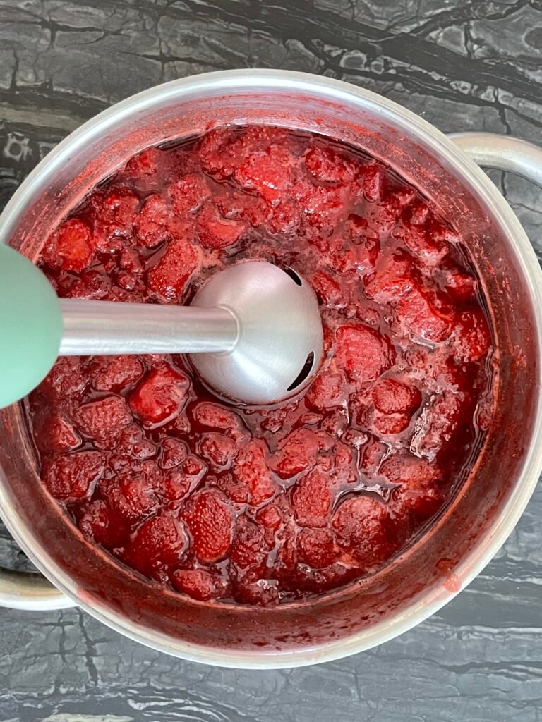 Immersion blender partially blended into a pot of chunky strawberry lime jam, smoothing part of the mixture while keeping some texture