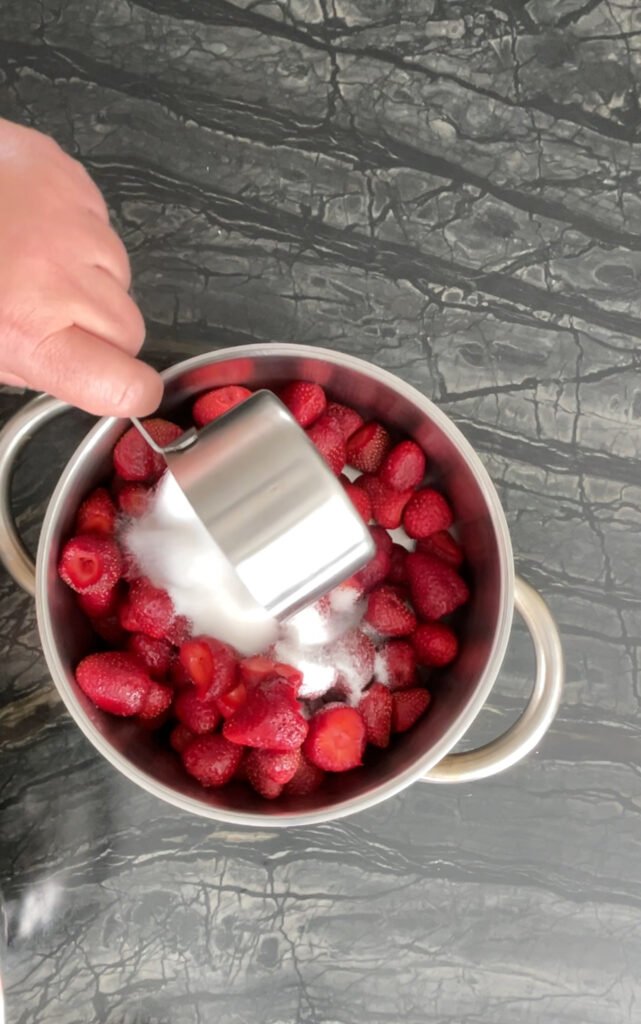 Hand pouring sugar from a metal measuring cup into a pot of hulled strawberries