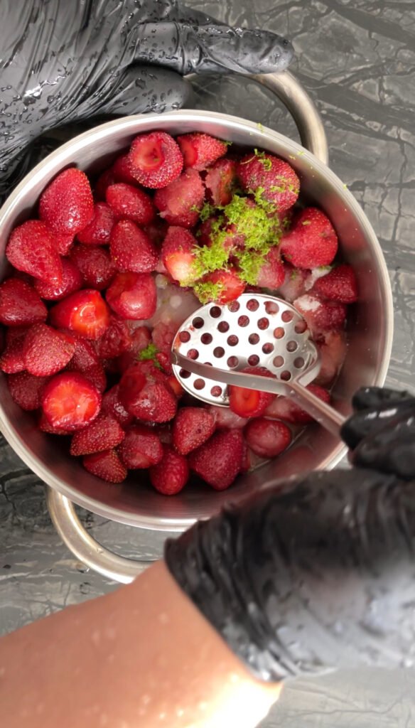 Gloved hand pressing a masher into a pot of strawberries topped with fresh lime zest