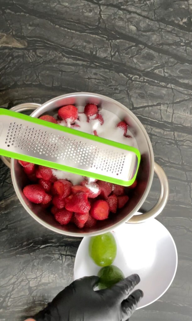 Hand zesting a lime with a microplane grater over a bowl, with a pot of strawberries in the background