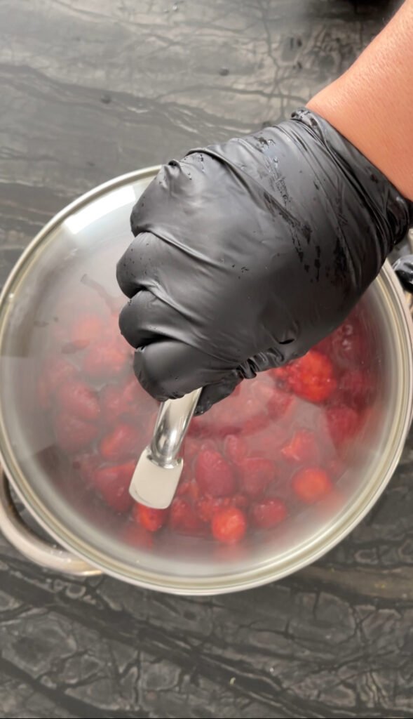 Gloved hand resting on a fogged glass lid covering a pot of strawberries, condensation visible from macerating