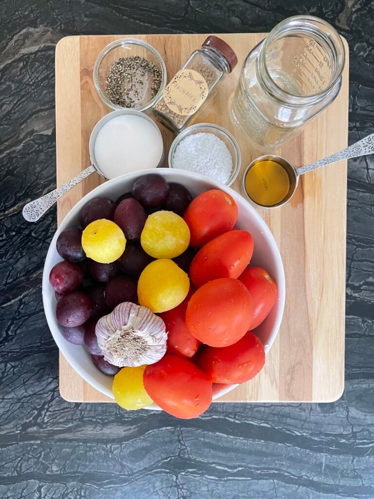 Overhead shot of raw ingredients for plum and tomato ketchup: tomatoes, purple and yellow plums, garlic bulb, and small bowls of spices and sugar on a wood cutting board.