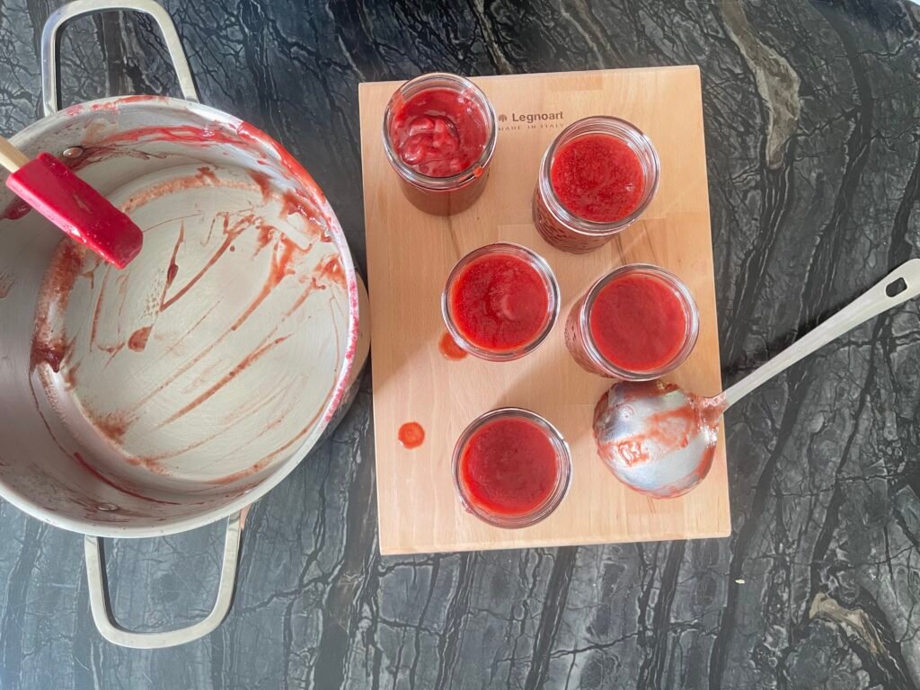 Overhead view of five glass canning jars filled with red ketchup arranged on a cutting board, next to jar lids and a bowl