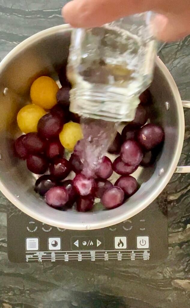 pot with the plums to help them break down as they cook. Hands pouring water from a glass measuring cup into a pot of whole purple and yellow plums.