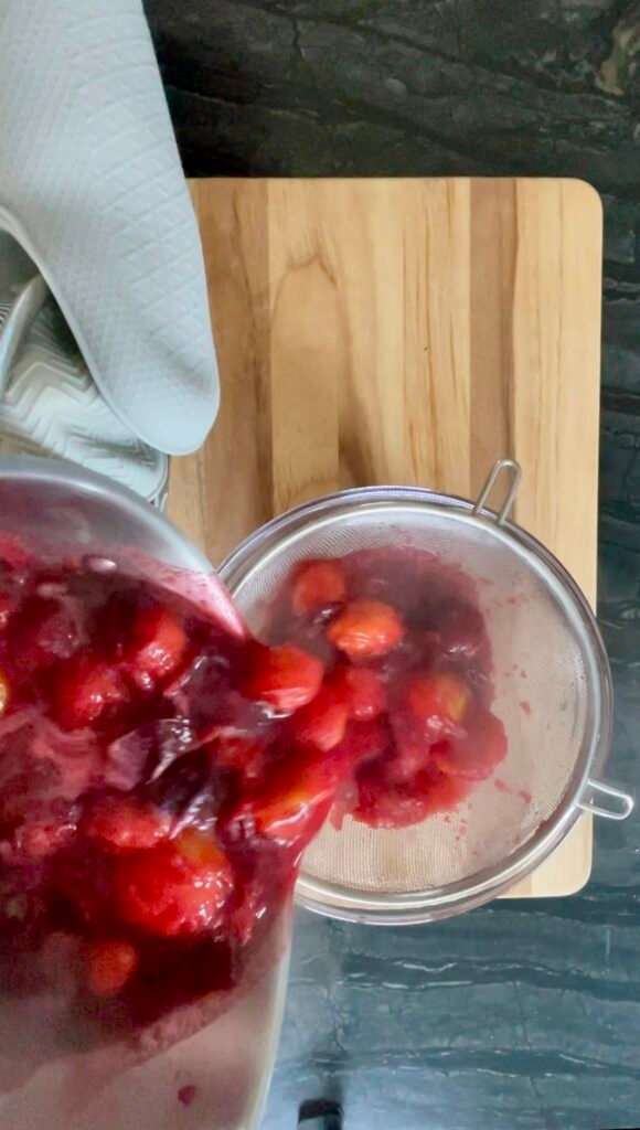 Gloved hands pouring cooked plums into a fine-mesh strainer set over a bowl.