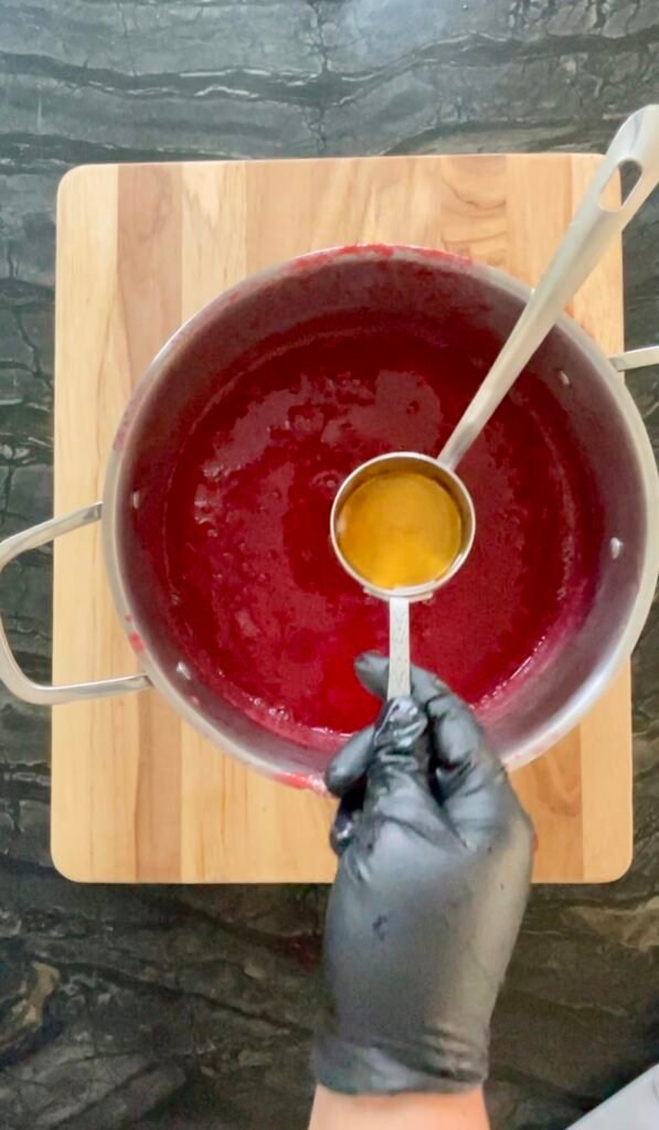 Gloved hand pouring a small amount of amber liquid from a measuring cup into a pot of red ketchup base