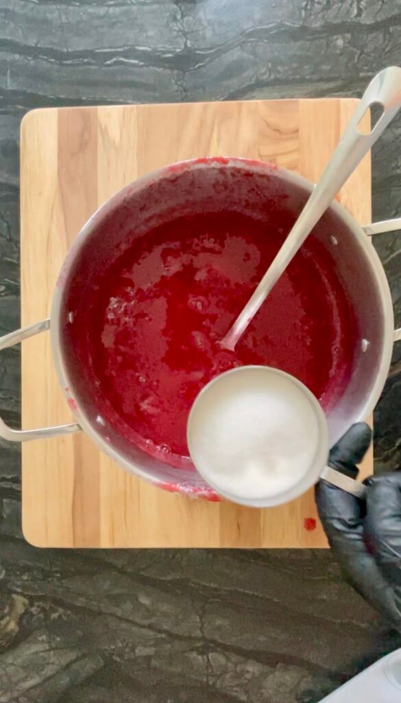 Overhead shot of a pot of red ketchup base with a spoon, a small white bowl of sugar beside it, ready to be added.