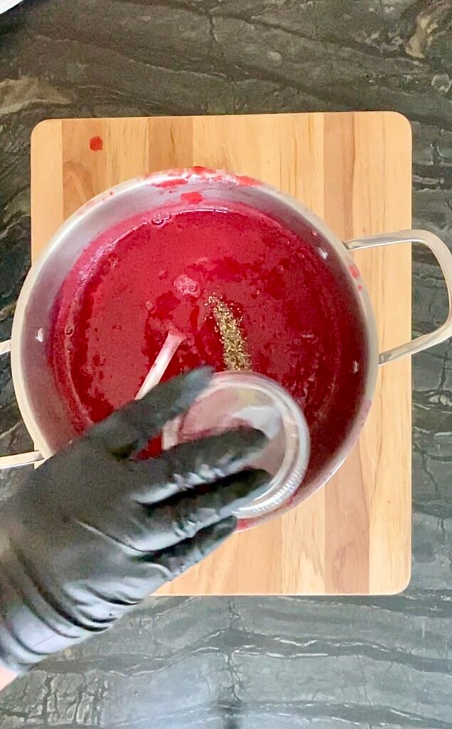 Gloved hand tipping ground black pepper from a small glass into a pot of red ketchup base, spoon resting in the pot.