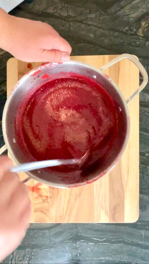 Overhead view of a pot of a red plum-tomato mixture, a spoon stirring in ground fenugreek