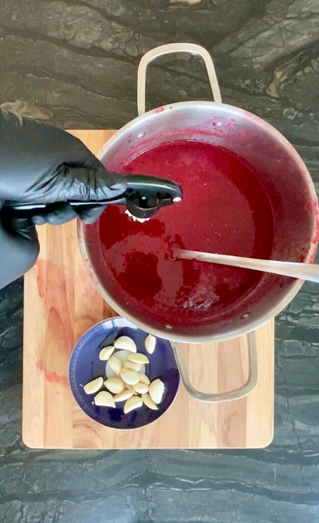 gloved hand stirring dark red ketchup base with a wooden spoon in a pot, a small blue bowl of pale sliced pieces resting on the cutting board nearby.