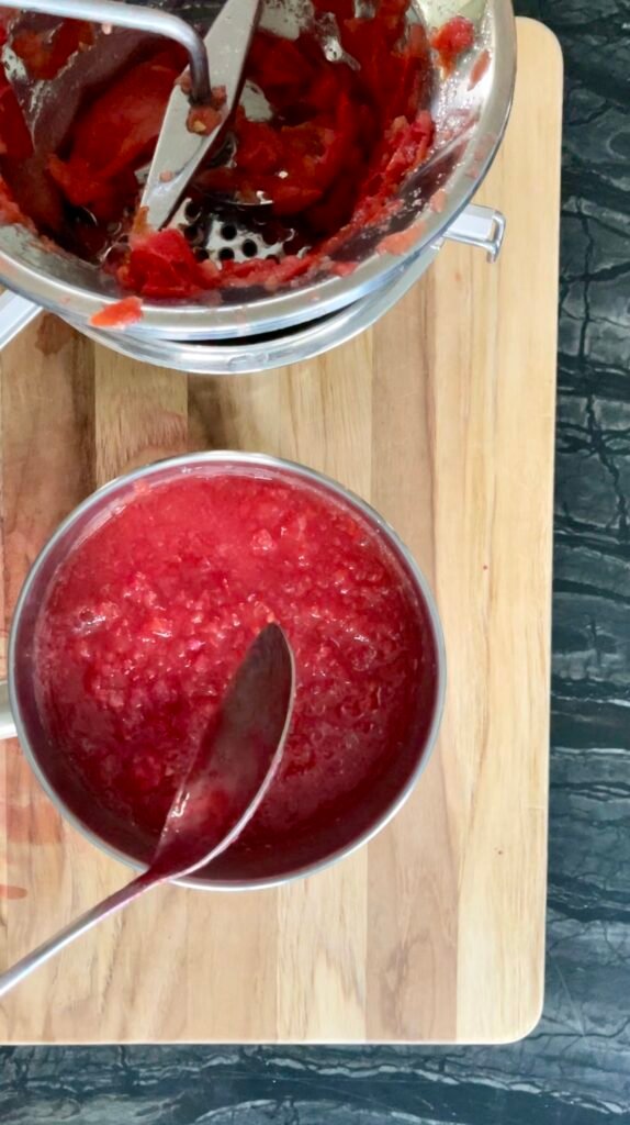 through a food mill to remove skins and seeds. Overhead view of a food mill with tomato skins and seeds left behind, a bowl of smooth tomato pulp underneath on a cutting board