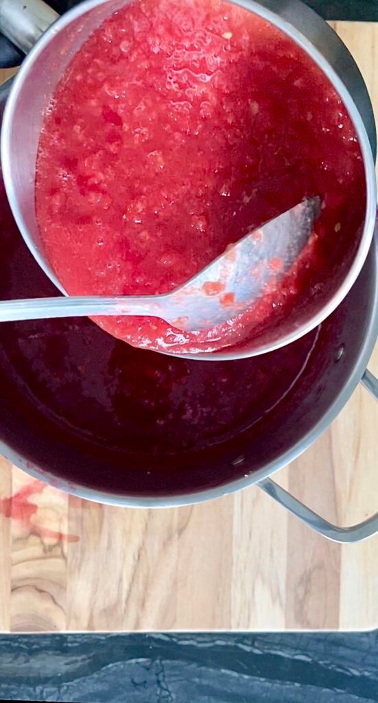 Overhead shot of a pot with thick red plum mixture, a spoon stirring in smooth tomato pulp poured in from a second pot.