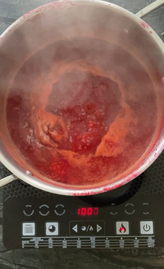 ketchup base until it starts to reduce. Overhead view of a pot of orange-red mixture simmering on an induction cooktop, steam rising, digital display visible on the burner.