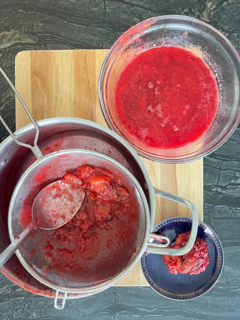 Gloved hands pressing cooked red plum pulp through a fine-mesh strainer set over a bowl.