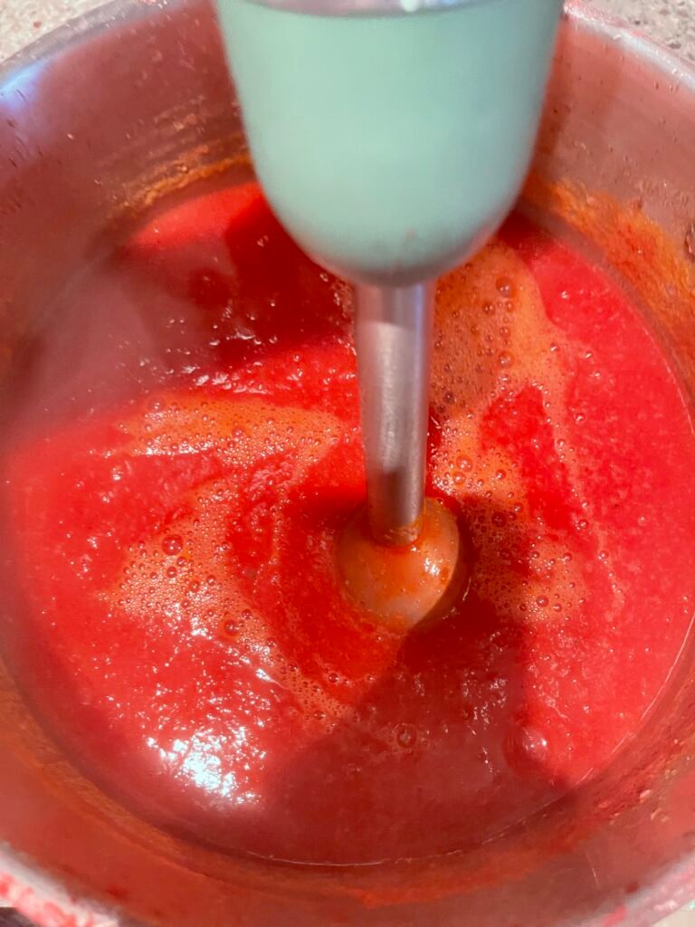 Close-up of a teal-handled immersion blender blending red ketchup mixture to a smooth, foamy consistency.