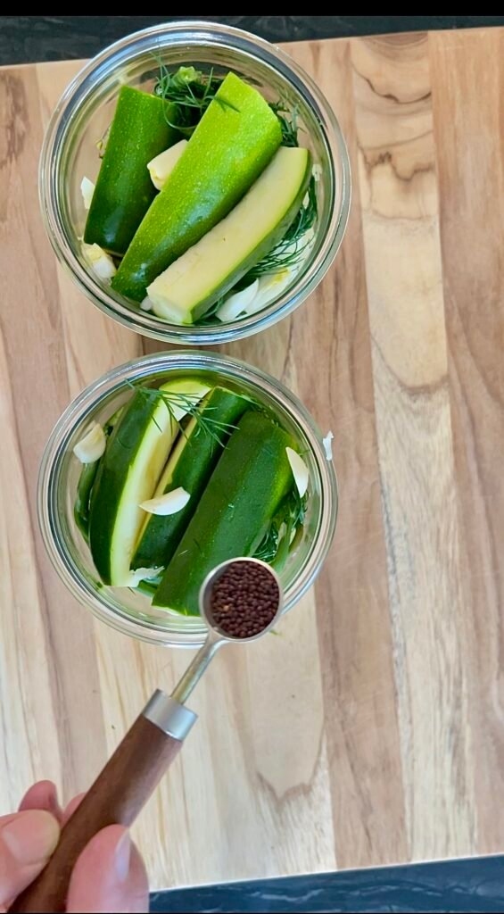 Spooning mustard seed into a jar packed with zucchini spears and dill