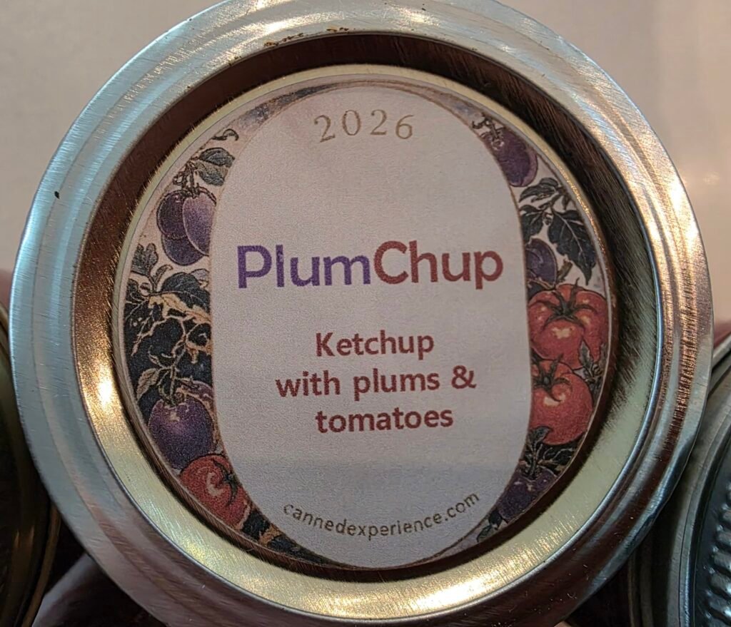 Overhead view of a canning jar lid with a custom "PlumChup" label reading "Ketchup with plums & tomatoes," dated 2026, with a tomato-and-plum vine illustration border and cannedexperience.com printed at the bottom.