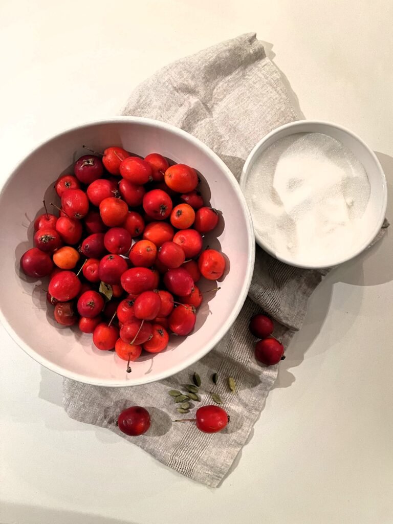 Bowl of whole red crab apples beside a small bowl of sugar and a few green cardamom pods on a linen cloth