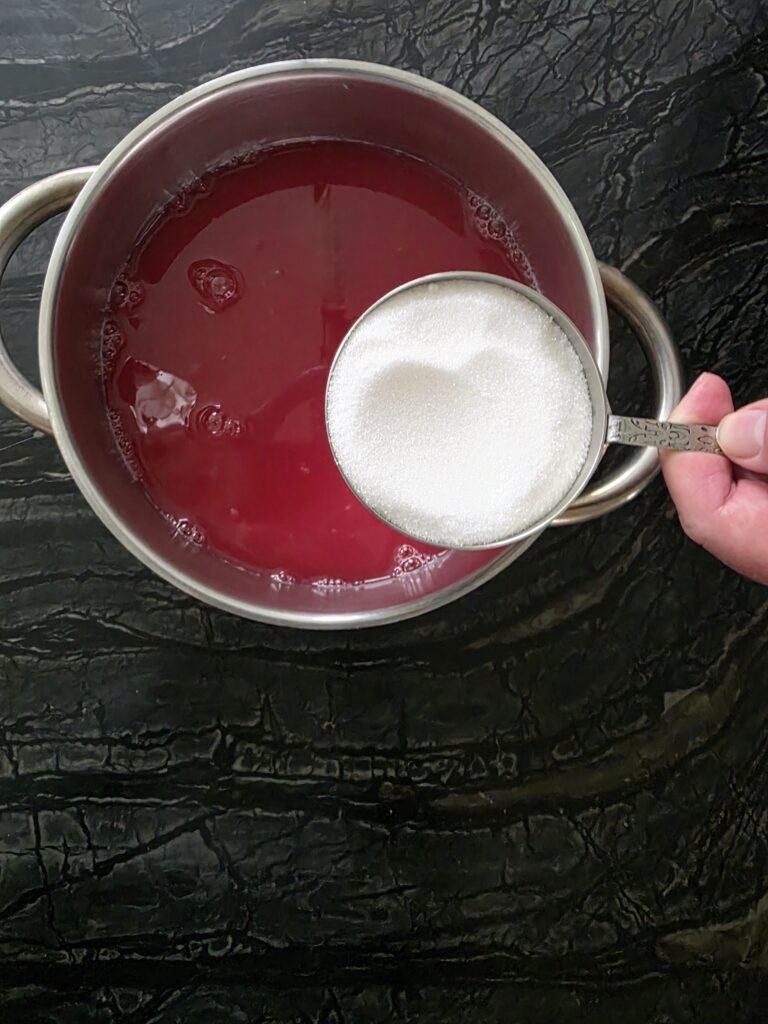 Measuring cup of white sugar held over a pot of red crab apple juice