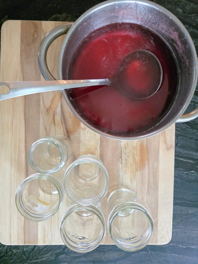 Pot of red crab apple jelly with a ladle on a cutting board next to five empty canning jars