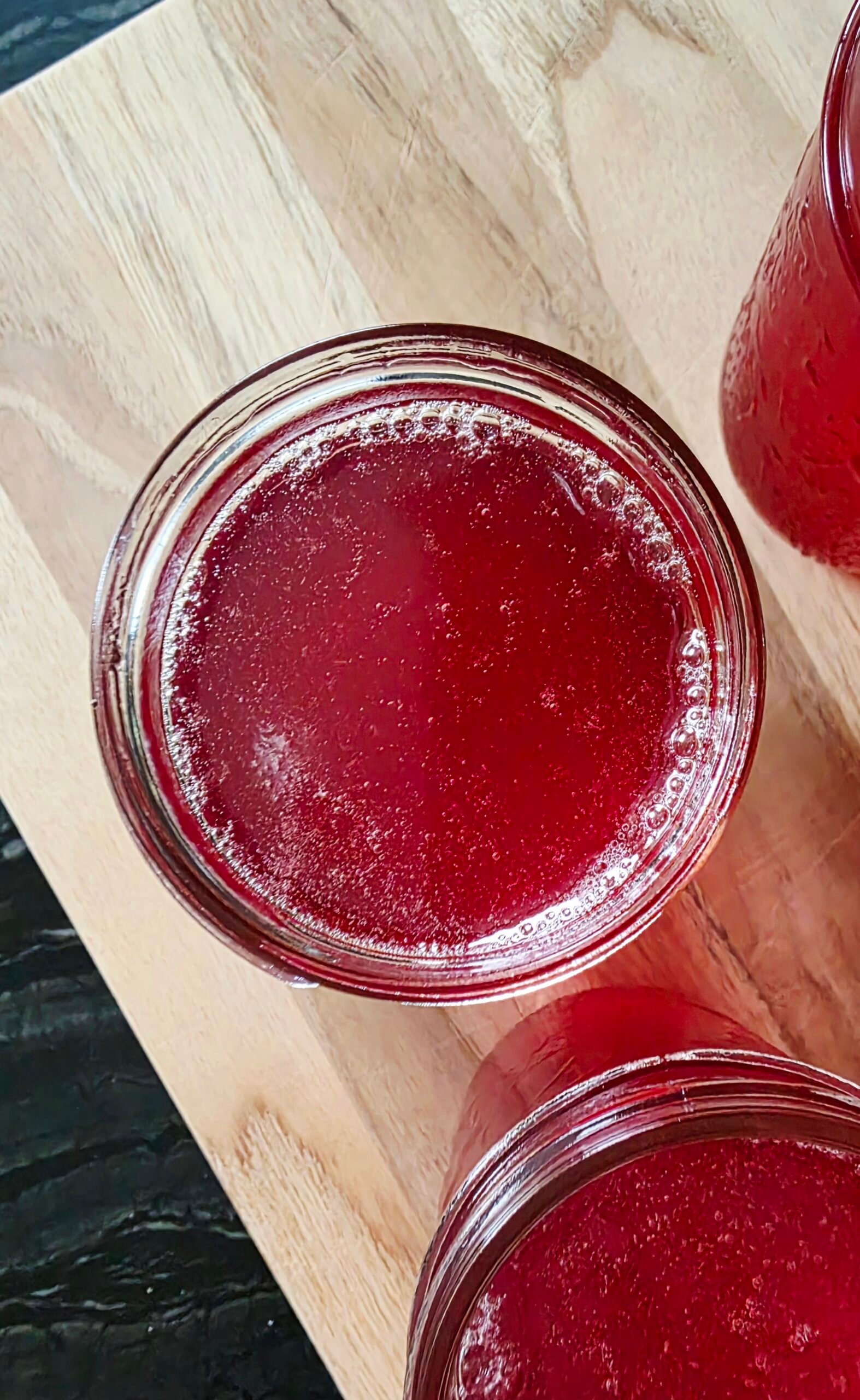 Top-down view of an open jar of red crab apple jelly with foam along the rim, on a wooden board with other jars
