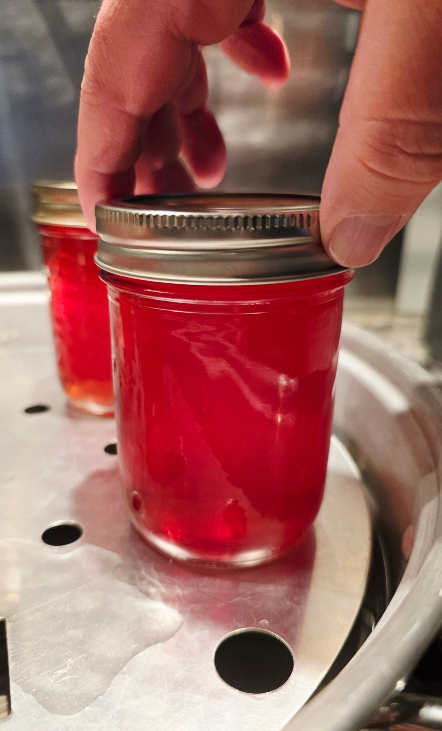 Hand on the lid of a jar of red crab apple jelly on a canner rack, with a second jar behind