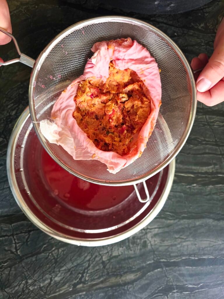 Cheesecloth bundle of crab apple pulp in a fine-mesh strainer over a bowl of red juice
