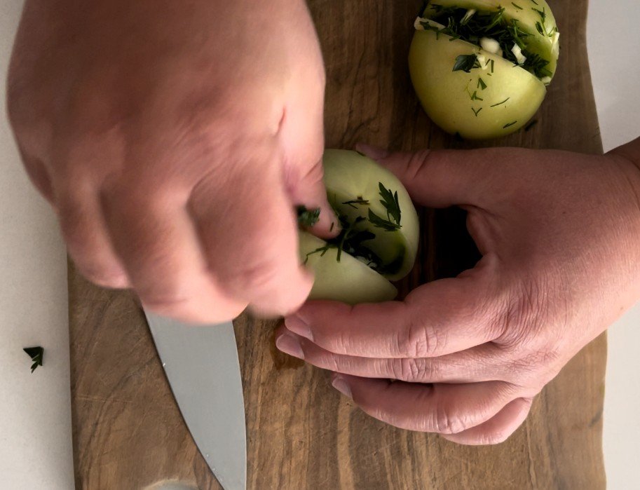 Hands pressing chopped herbs and garlic into a cut green tomato on a wooden board