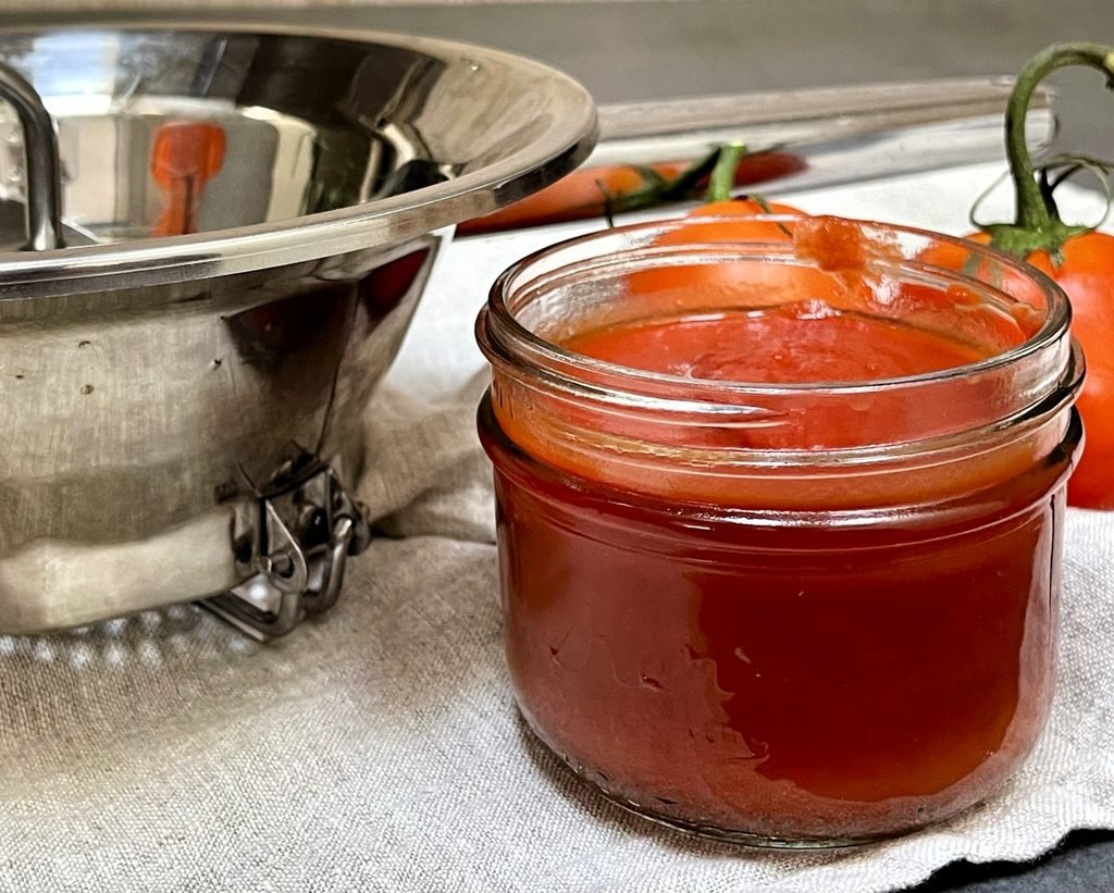 Small jar of smooth tomato sauce beside a stainless steel food mill and fresh tomatoes