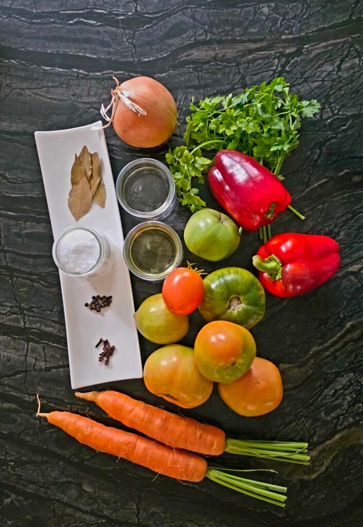 Onion, parsley, red and orange peppers, tomatoes, carrots, bay leaves, cloves, and salt arranged on a dark surface.