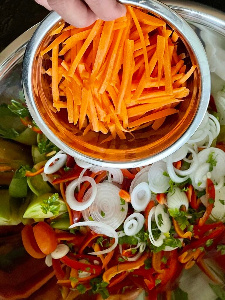 Carrot matchsticks being poured over a bowl of onion rings, peppers, and tomatoes.