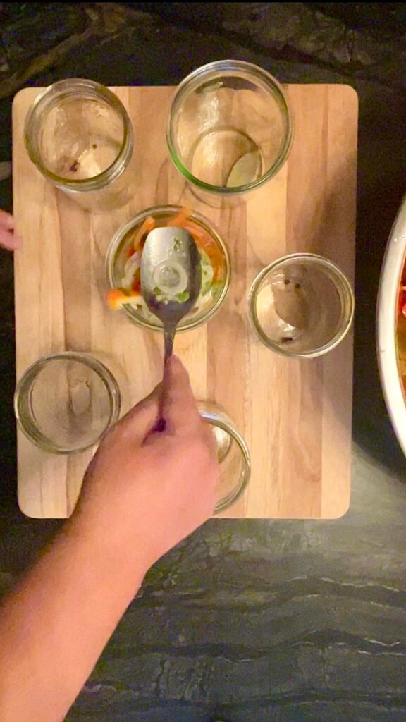 A spoon pressing vegetables into a glass jar on a wooden board, with other empty jars nearby