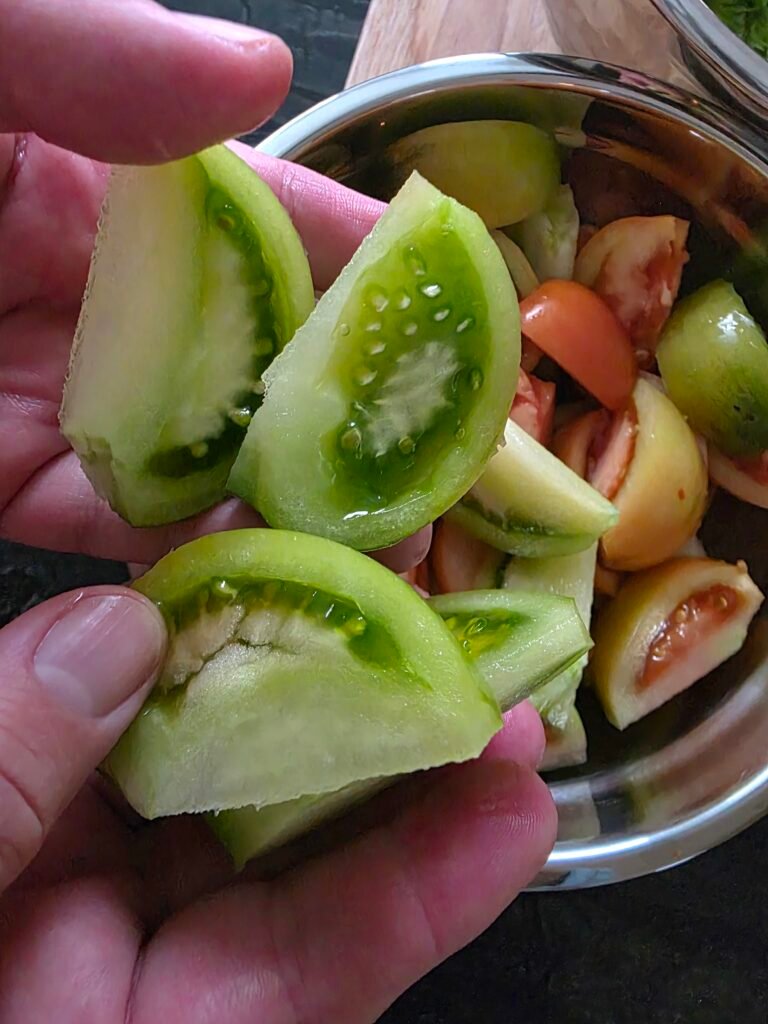 A hand holding green tomato wedges over a bowl of sliced tomatoes.