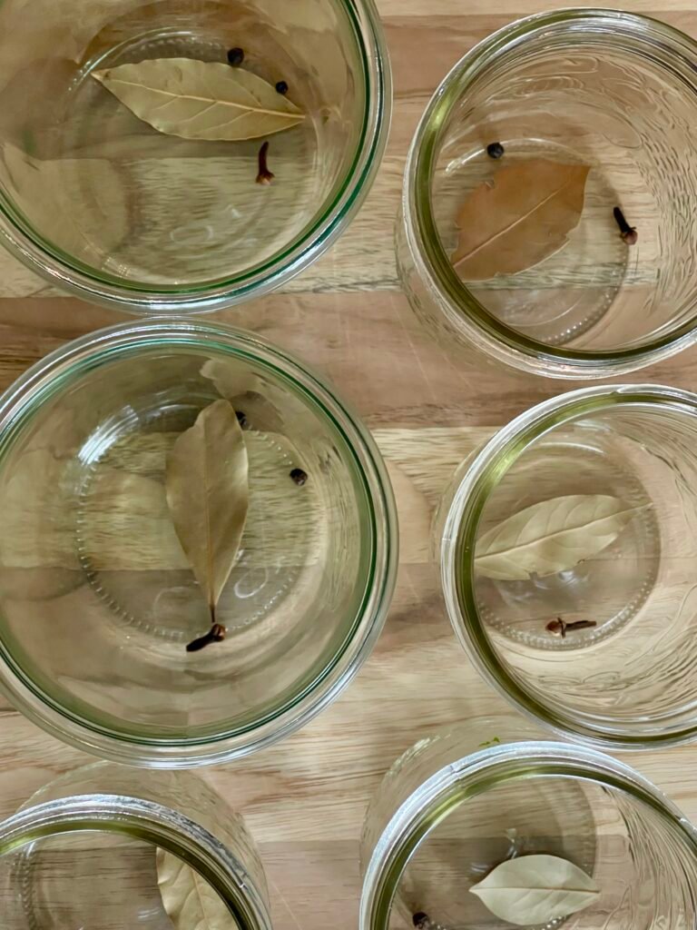 Empty glass jars, each with a bay leaf, peppercorns and a clove.